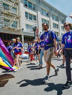a group of PSU affiliates walk in the Portland Pride parade in matching purple shirts. A white queer couple holds hands and waves