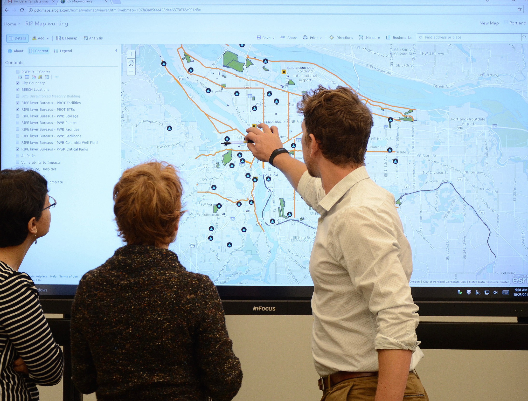 Three people face a large screen with a map of Portland on it. Person furthest right is touching screen to expand the view