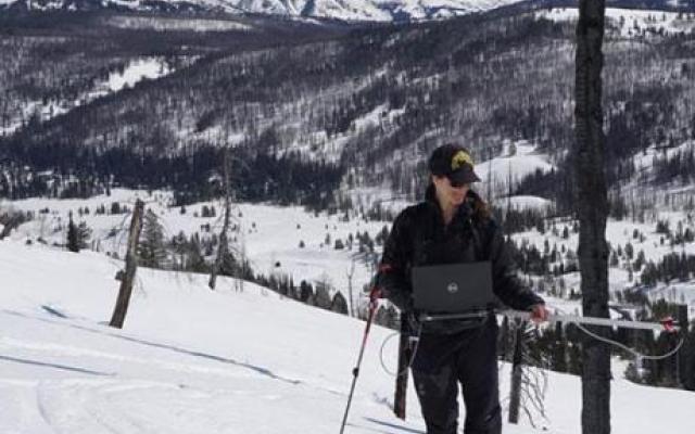 Kelly Gleason stands with a laptop in the snow. Mountains are visible behind her.