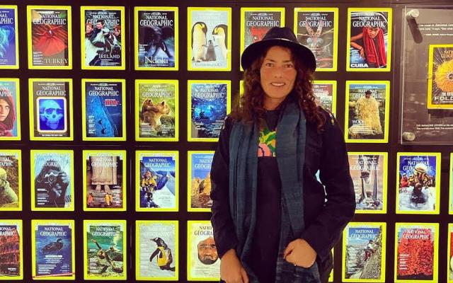Film director Katie Conlon stands in front of a wall of National Geographic magazine covers. She wears a dark hat and blue scarf, and faces the camera