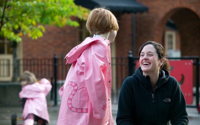 A young child in a pink raincoat has her back turned to us as she is facing a smiling brown-haired adult woman.