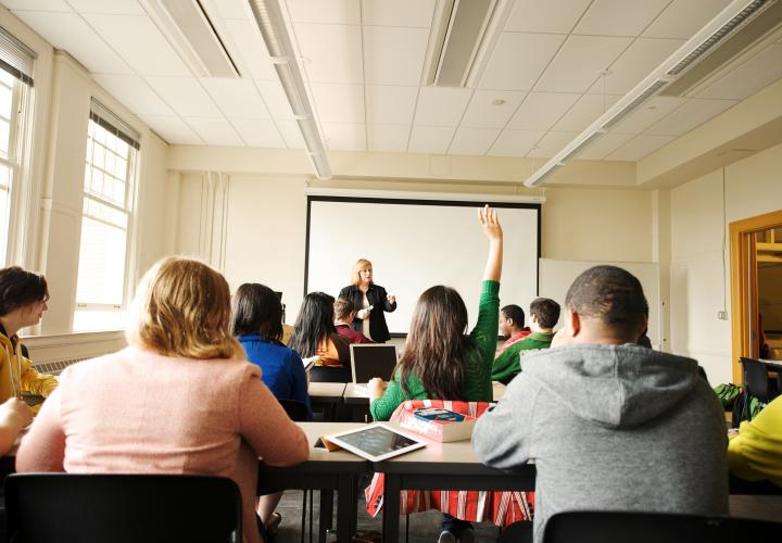 The backs of students' heads as they sit facing a standing teacher. One student has a hand raised.