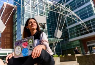 Student sitting with a laptop in front of a building with many glass windows.