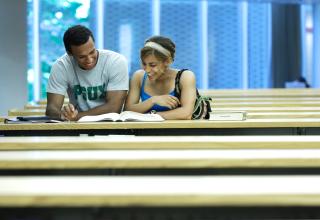 Two students standing among the stacks of the library laughing at an open book