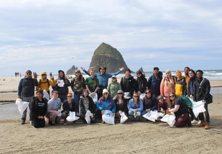 A group of people is shown standing in front of the ocean and a large rock. The students in the front row are holding trash bags.