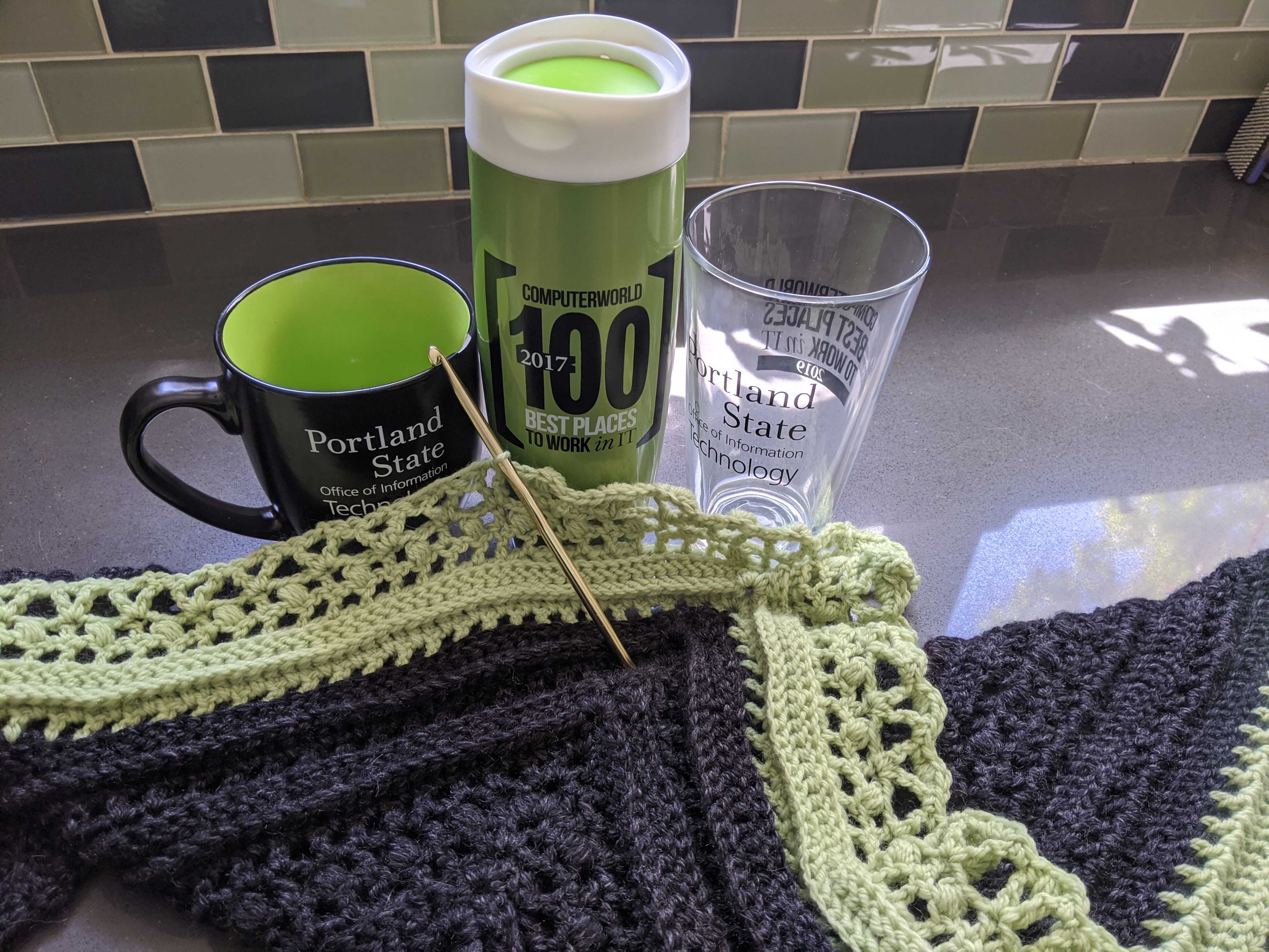 Three mugs celebrating our awards for Best Place to Work in IT, sitting on a countertop next to a shawl crocheted in the PSU colors.