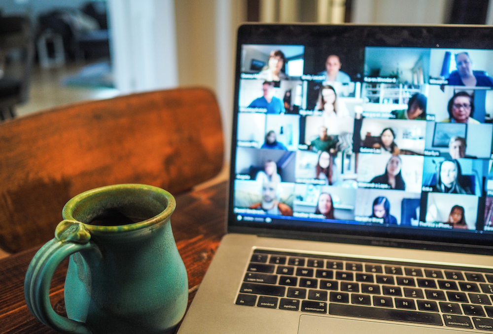 Open laptop on a table with a mug in foreground. A multi-person Zoom meeting is shown on the laptop's screen.