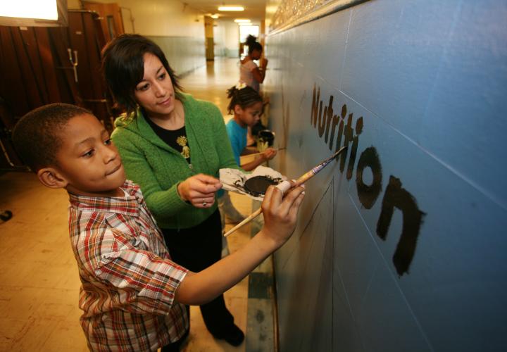 Faculty member and a grade school student paint the word nutrition on hallway wall