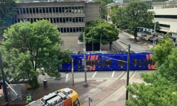 A photo of a blue MAX and an orange streetcar surrounded by trees and buildings taken from above